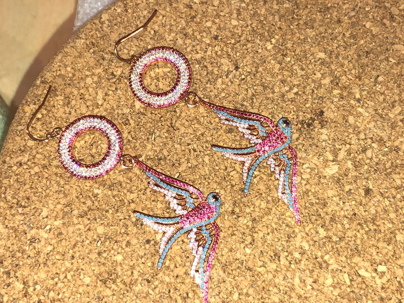 Puede incluir: Un par de pendientes de tono dorado con un dise&ntilde;o circular y un p&aacute;jaro golondrina colgante. Los pendientes est&aacute;n adornados con pedrer&iacute;a rosa, azul y blanca.