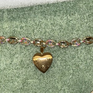 May include: A gold chain bracelet with a heart-shaped locket pendant. The chain is made of oval links with rainbow-colored stones. The locket has a floral design.