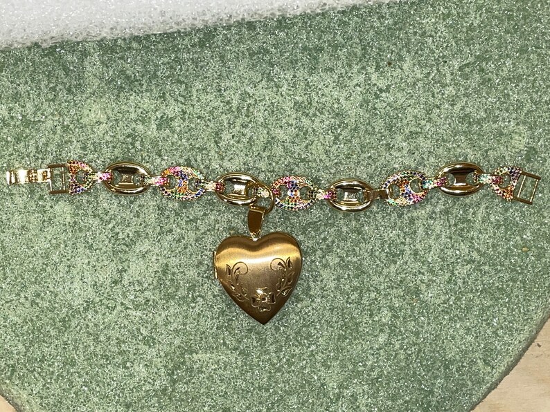 May include: A gold chain bracelet with a heart-shaped locket pendant. The chain is made of oval links with rainbow-colored stones. The locket has a floral design.