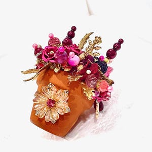 May include: A small terracotta flower pot decorated with a variety of pink, red, and gold embellishments. The pot is topped with a large, gold, rhinestone flower.