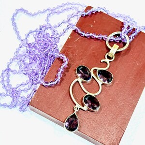 May include: A silver necklace with a pendant featuring a cluster of amethyst gemstones in a delicate, abstract design. The necklace is made with a strand of light purple beads.
