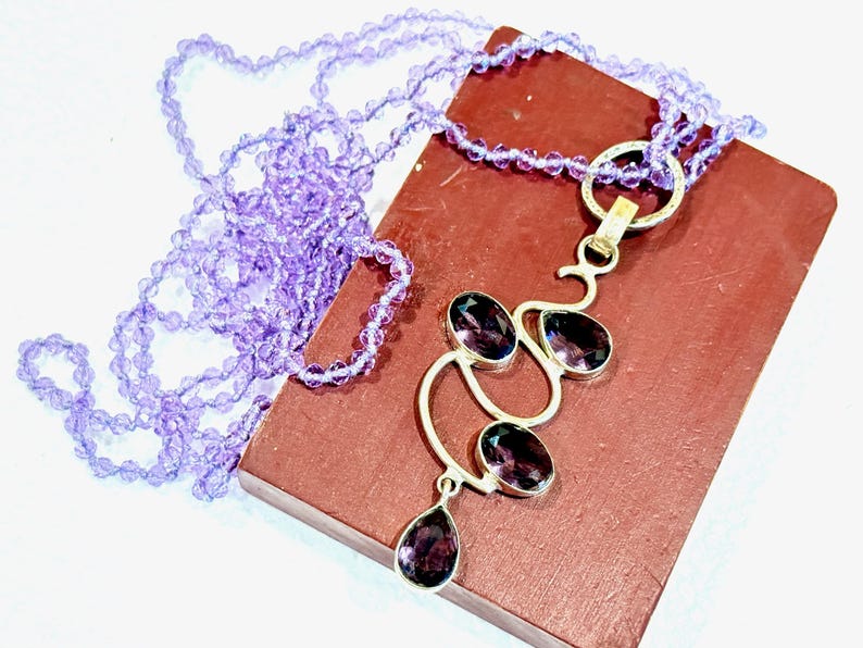 May include: A silver necklace with a pendant featuring a cluster of amethyst gemstones in a delicate, abstract design. The necklace is made with a strand of light purple beads.