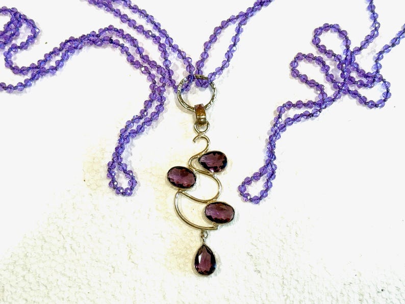 May include: A purple amethyst gemstone necklace with a pendant featuring a gold swirl design and multiple amethyst stones.