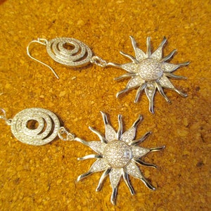 May include: A pair of silver earrings with a sunburst design and a circle pendant. The sunburst design is covered in clear rhinestones.