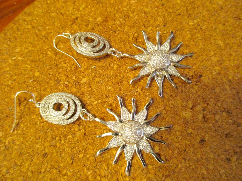 May include: A pair of silver earrings with a sunburst design and a circle pendant. The sunburst design is covered in clear rhinestones.