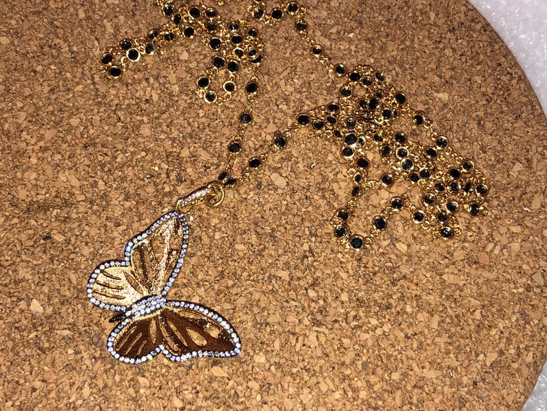 May include: A gold chain necklace with a butterfly pendant. The chain is made of small gold links with black beads. The butterfly pendant is detailed with gold and white accents.