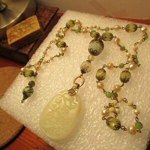 Puede incluir: Un collar con un colgante de jade verde y blanco con un dise&ntilde;o tallado. El collar est&aacute; hecho con cuentas de vidrio verde y blanco y una cadena de oro.