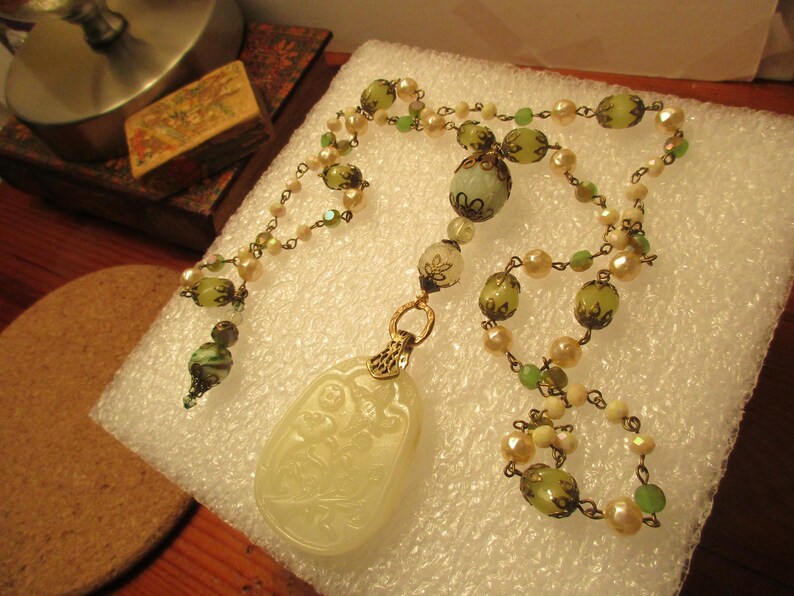 Puede incluir: Un collar con un colgante de jade verde y blanco con un dise&ntilde;o tallado. El collar est&aacute; hecho con cuentas de vidrio verde y blanco y una cadena de oro.