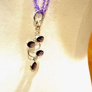 May include: A silver necklace with a pendant featuring a cluster of four amethyst stones. The pendant is shaped like a stylized tree branch with the stones set in the branches. The necklace is made of a double strand of small purple beads.