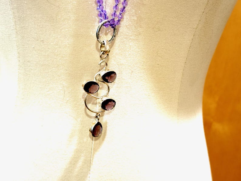 May include: A silver necklace with a pendant featuring a cluster of four amethyst stones. The pendant is shaped like a stylized tree branch with the stones set in the branches. The necklace is made of a double strand of small purple beads.
