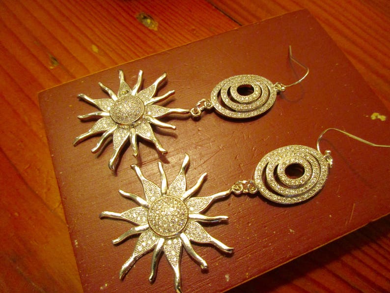 May include: Two pairs of silver earrings with a sunburst and circle design. The earrings are adorned with sparkling rhinestones.