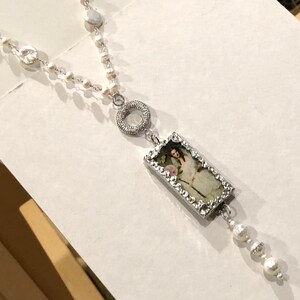 Pu&ograve; includere: Una collana d'argento con una catena di perle bianche e un ciondolo rettangolare con un'immagine vintage di una donna. Il ciondolo &egrave; circondato da una cornice d'argento con piccole pietre trasparenti. La collana ha anche un ciondolo circolare d'argento con pietre trasparenti.