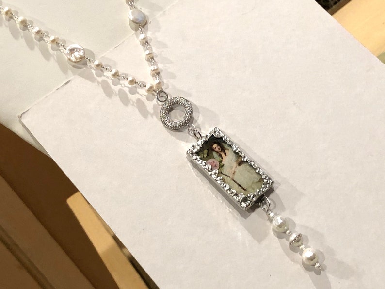 Pu&ograve; includere: Una collana d'argento con una catena di perle bianche e un ciondolo rettangolare con un'immagine vintage di una donna. Il ciondolo &egrave; circondato da una cornice d'argento con piccole pietre trasparenti. La collana ha anche un ciondolo circolare d'argento con pietre trasparenti.
