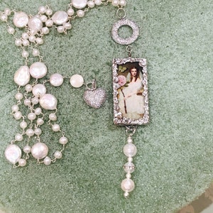 Pu&ograve; includere: Una lunga collana a catena d'argento con perle bianche e un ciondolo rettangolare con un ritratto vintage di una donna in un abito bianco che tiene una rosa rosa. Il ciondolo &egrave; circondato da una cornice d'argento con un design decorativo. La collana include anche un ciondolo a forma di cuore d'argento con strass trasparenti e un ciondolo circolare d'argento.