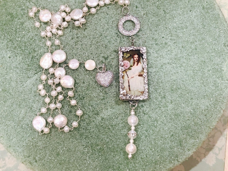 Pu&ograve; includere: Una lunga collana a catena d'argento con perle bianche e un ciondolo rettangolare con un ritratto vintage di una donna in un abito bianco che tiene una rosa rosa. Il ciondolo &egrave; circondato da una cornice d'argento con un design decorativo. La collana include anche un ciondolo a forma di cuore d'argento con strass trasparenti e un ciondolo circolare d'argento.