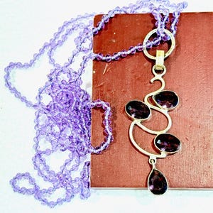 May include: A silver necklace with a pendant featuring a cluster of amethyst gemstones in a swirling design. The necklace is paired with a strand of purple beads.