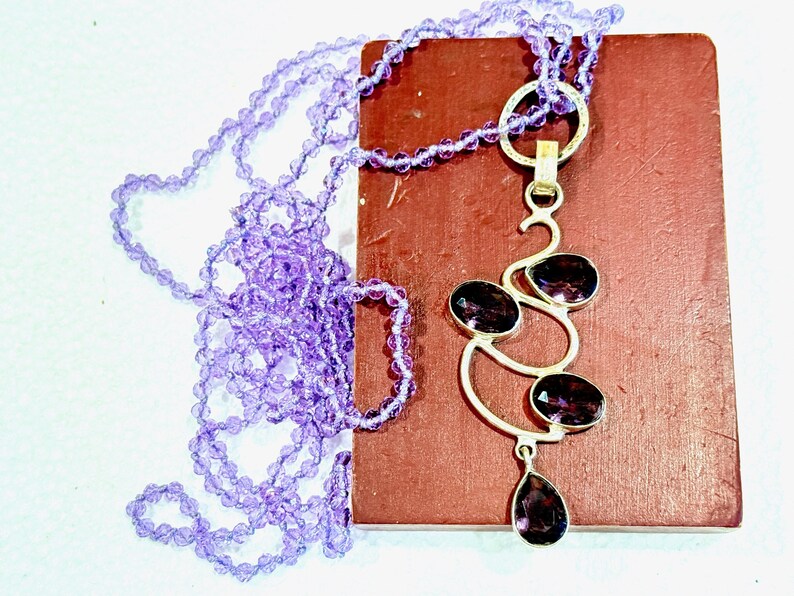 May include: A silver necklace with a pendant featuring a cluster of amethyst gemstones in a swirling design. The necklace is paired with a strand of purple beads.