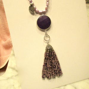 May include: A purple and silver necklace with a large round pendant and a tassel. The pendant is made of a dark purple stone and the tassel is made of small purple beads.