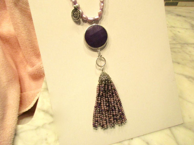 May include: A purple and silver necklace with a large round pendant and a tassel. The pendant is made of a dark purple stone and the tassel is made of small purple beads.