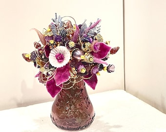 May include: A purple glass vase with a floral design holds a bouquet of purple, white, and gold flowers. The flowers are made of fabric, beads, and other decorative elements.