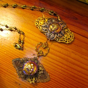 Może przedstawiać: Złoty naszyjnik z dwoma ozdobnymi zawieszkami. Zawieszki mają kwiatowy wz&oacute;r z perłowym guzikiem w środku. Naszyjnik wykonany jest z łańcuszka z małymi złotymi koralikami.