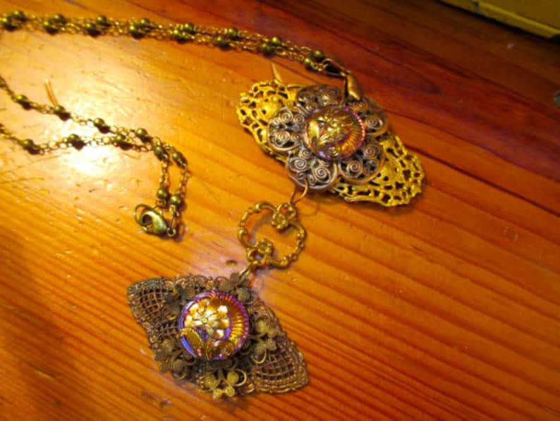 Może przedstawiać: Złoty naszyjnik z dwoma ozdobnymi zawieszkami. Zawieszki mają kwiatowy wz&oacute;r z perłowym guzikiem w środku. Naszyjnik wykonany jest z łańcuszka z małymi złotymi koralikami.