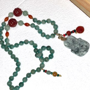 Puede incluir: Un collar de jade verde con un colgante de jade tallado y cuentas rojas y naranjas.