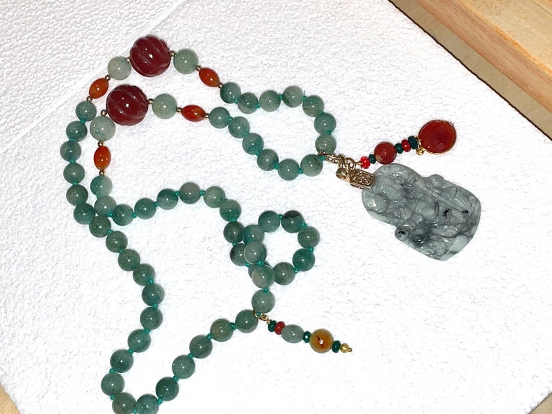 Puede incluir: Un collar de jade verde con un colgante de jade tallado y cuentas rojas y naranjas.