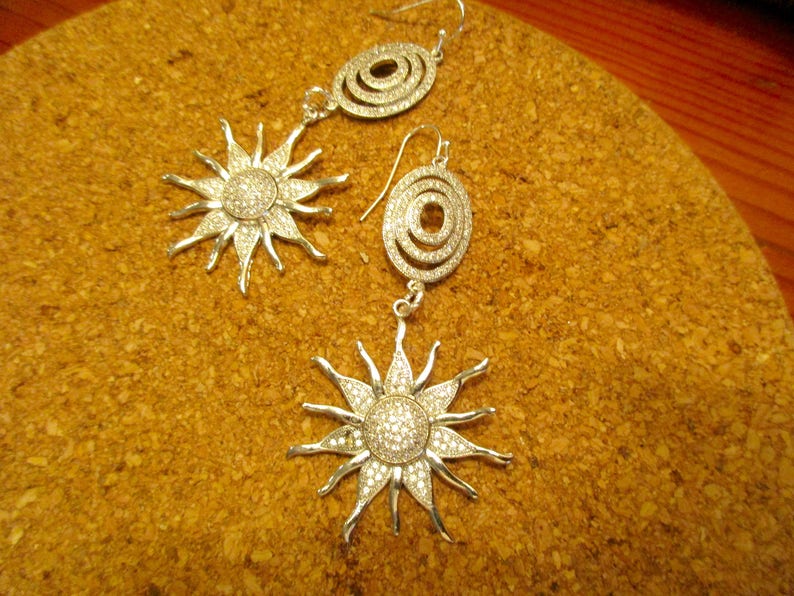 May include: A pair of silver earrings with a sunburst design and a circular design. The earrings are set with clear crystals.