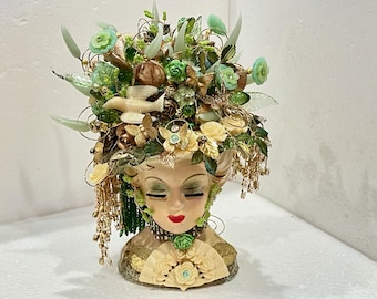 May include: A ceramic bust of a woman with a face and neck, wearing a crown of green and gold leaves, flowers, and beads. The crown is adorned with butterflies and other decorative elements.