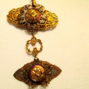 Może przedstawiać: Złoty naszyjnik z wisiorem, na kt&oacute;rym znajdują się dwa tęczowe szklane kaboszony osadzone w ozdobnym metalowym filigranie. Wisiorek ma kwiatowy wz&oacute;r i jest zawieszony na łańcuszku.