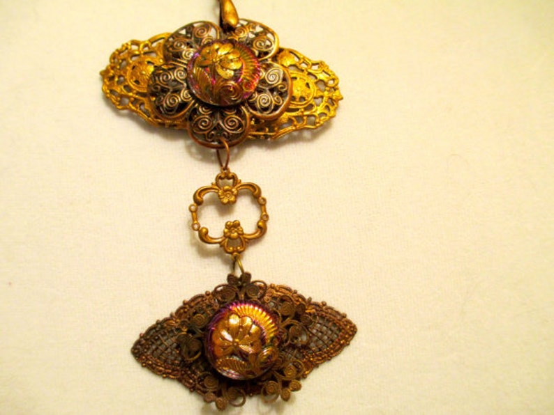 Może przedstawiać: Złoty naszyjnik z wisiorem, na kt&oacute;rym znajdują się dwa tęczowe szklane kaboszony osadzone w ozdobnym metalowym filigranie. Wisiorek ma kwiatowy wz&oacute;r i jest zawieszony na łańcuszku.