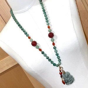 Puede incluir: Un collar de jade verde con cuentas rojas y naranjas. El collar tiene un colgante de jade en forma de amuleto de la suerte chino.