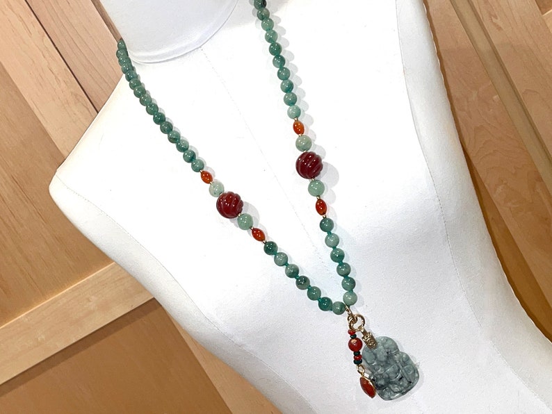 Puede incluir: Un collar de jade verde con cuentas rojas y naranjas. El collar tiene un colgante de jade en forma de amuleto de la suerte chino.