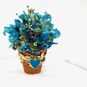 May include: A small terracotta pot with a gold trim and a blue heart is filled with a blue and gold beaded flower arrangement.