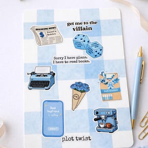 Könnte beinhalten: Eine Sammlung von Aufklebern auf einem blau-weiß karierten Hintergrund. Die Aufkleber zeigen verschiedene Designs, darunter eine Zeitung, Würfel, eine Schreibmaschine, einen Blumenstrauß, eine Kaffeemaschine und Text.