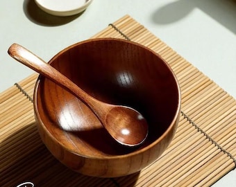 May include: A wooden bowl and spoon set. The bowl and spoon are made of a dark brown wood. The spoon rests inside the bowl. A small, off-white ceramic dish is in the background. The word "Grainism" is written in cursive at the bottom.