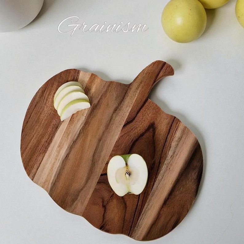 May include: A wooden cutting board shaped like an apple, with a handle. The board has a natural wood grain pattern and contains sliced apples. The word "Grainism" is written in cursive on the top. The overall theme is kitchen and food preparation.