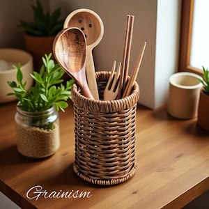 Puede incluir: Un soporte de utensilios tejido de mimbre lleno de cucharas de madera, un tenedor y palillos. El soporte es marrón y está sobre una superficie de madera. La palabra "Grainism" está escrita en cursiva en la parte inferior.