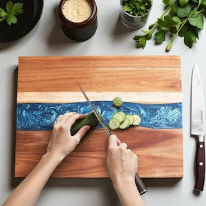 May include: A wooden chopping board with a blue resin river design. A person is slicing a cucumber with a stainless steel knife. The board is rectangular and has a light wood border. Sliced cucumber pieces are visible on the board.