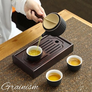 以下が含まれることがあります： 木製のハンドルが付いた黒と金のティーセットが、小さな白黒のティーカップに紅茶を注いでいます。 ティーセットは、紅茶の入った他の2つのティーカップが入った濃い木製のトレイの上にあります。 「Grainism」という言葉が左下にあります。