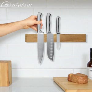 May include: Three stainless steel kitchen knives of varying sizes are displayed on a wooden magnetic knife holder. The knives are silver with black handles. The wooden holder is mounted on a white tiled wall. The brand name "Grainism" is in the top left corner.