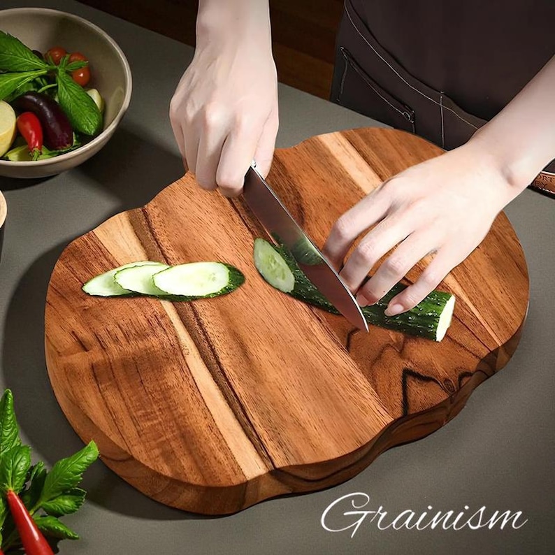 May include: A wooden cutting board with a wavy edge is used to slice a cucumber. The board has a natural wood grain pattern and is being used with a stainless steel knife. Sliced cucumber pieces are visible on the board. The word "Grainism" is written on the board.
