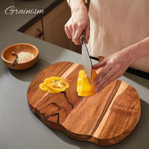 May include: A wooden cutting board with a unique shape, featuring a person slicing a yellow bell pepper. The board has a natural wood grain pattern and is accompanied by a small wooden bowl. The text "Grainism" is visible in the upper left corner.