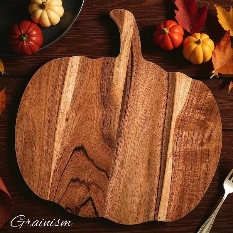 May include: A wooden pumpkin-shaped cutting board with a handle. The board is made of light and dark brown wood. Miniature pumpkins in red, orange, and yellow are arranged around the board, along with fall leaves. The word "Grainism" is at the bottom.