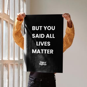 May include: A black poster with the text "BUT YOU SAID ALL LIVES MATTER" in white. The poster is held up in front of a person. The person is wearing a yellow knit sweater and black jeans. The words "Signs of dissent" are at the bottom.