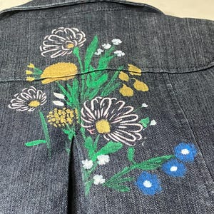 May include: A dark gray denim jacket featuring a hand-painted floral design on the back. The artwork displays white daisies with yellow centers, green stems and leaves, small yellow clusters, and two blue flowers with white centers near the bottom.