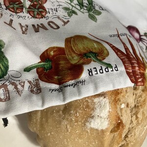 May include: A loaf of bread partially covered by a white kitchen towel. The towel features illustrations of tomatoes, peppers, and other vegetables, with text that reads "TOMATO" and "PEPPER". The bread has a light brown crust.
