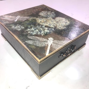 May include: A rectangular decorative box with a dark gray body and a lid featuring a dragonfly and floral design. The lid has a distressed, vintage look with gold trim. The box has a metal handle and rests on small feet.