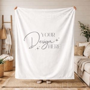 Puede incluir: Una manta blanca grande, sostenida por una persona, con el texto "YOUR Design HERE" en escritura gris. La manta parece suave y afelpada. El fondo incluye una cesta de mimbre, una escalera de madera y un sofá. La manta mide aproximadamente 200 cm x 150 cm.